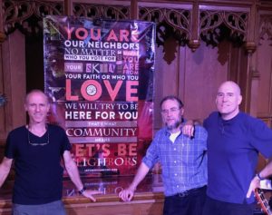 Members of the band Abraham Jam with a Let's Be Neighbors banner, hanging in the front of a chapel at Westminster College.